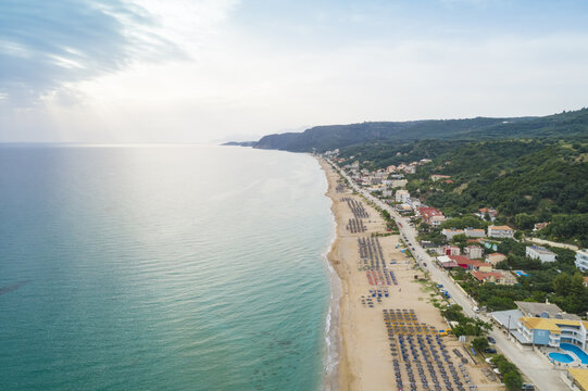 Aerial view of the sun-kissed beach lined with neat rows of umbrellas, buildings nestled against lush green hills, and the turquoise sea stretching to the horizon, Paralia, Preveza, Greece.