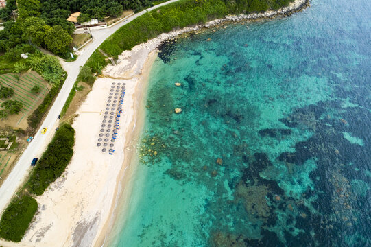 Aerial view of the golden sands meeting the turquoise sea, dotted with sun umbrellas and boats, framed by lush greenery, Paralia, Preveza, Greece.