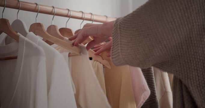 Close up of woman hand running across hanging clothes and taking beige sweater from rack. Female choosing outfit in bedroom at home.