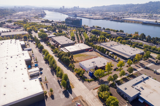 Aerial view of industrial buildings nestled among vibrant trees alongside a wide river reflecting the sky's soft hues, Quantum Assembly, Portland, United States.
