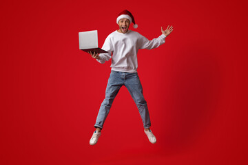 A young person wearing a Christmas hat jumps in excitement while holding a laptop. The vibrant red...