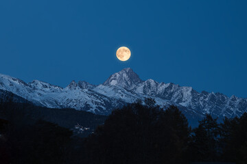 View of a luminous full moon ascends over the snow-capped, jagged peaks, casting a serene glow on the landscape, Evolene, Valais, Switzerland.