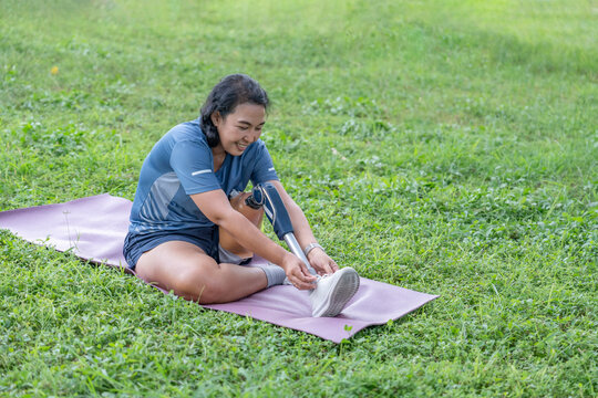 asian woman with prosthetic leg in sportswear sitting on yoga mat and tying shoelace in the park
