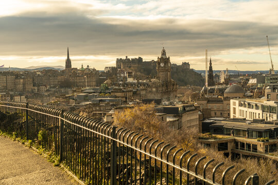 View of the golden cityscape with historic buildings and monuments under a cloudy sky, seen from behind a black metal fence, Edinburgh, Scotland, United Kingdom.