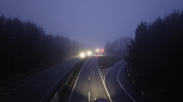 View from an overpass onto the two lane D1 highway with cars driving through dense fog at night, featuring blurred headlights and taillights creating atmospheric low visibility traffic conditions
