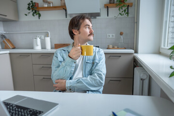 Calm reflective man holding mug of coffee in home kitchen, looking away deep in thought. Pensive young man with cup in hand taking break at table, gazing out window. Inner dialogue, decision making.