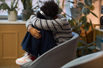 Black teenager sitting curled up in chair wearing headphones, hugging knees to chest, appearing withdrawn in school office setting, plants and window in background, school principal context
