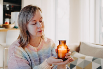 A woman gently holds a lit candle jar with both hands in a bright domestic setting. Mindfulness practice, meditation at home, finding stillness, intentional living.