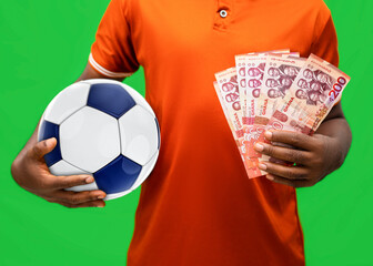 An individual in an orange shirt holds a blue and white soccer ball under one arm while displaying a fan of Ghanaian 200 Cedi banknotes in their other hand against a vibrant green background.