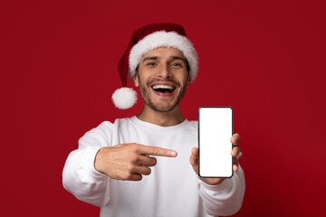 A man wearing a Santa hat smiles widely while pointing at his smartphone with a blank screen. The...