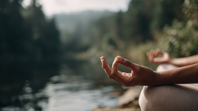 A person is meditating outdoors by a body of water with hands in a mudra gesture. This scene takes place in a natural area with soft light, creating a peaceful setting