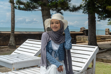 A woman wearing a hijab and hat is enjoying a vacation at a hotel with a beautiful beach view.