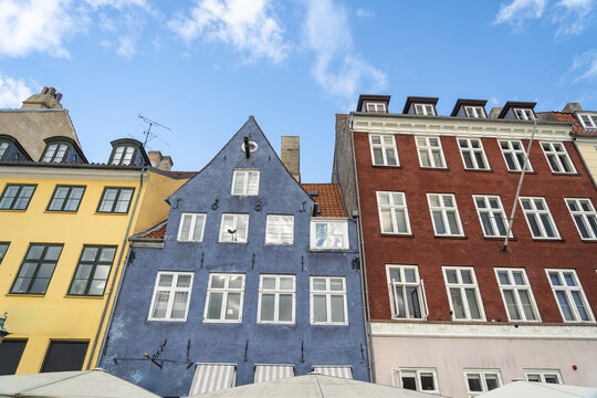 View of vibrant, colorful buildings stand shoulder to shoulder, their facades a patchwork of yellow, blue, and red under a sky brushed with clouds, Nyhavn, Copenhagen, Denmark.