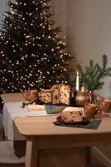 Festive Christmas holiday table setting, wooden table with food, panettone, tea cups, linen napkins, pine branch and candlelight, blurred Christmas tree lights on background, lifestyle home interior