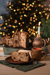 Sliced piece of panettone cake on plate, tea cups and teapot, candle light and green linen napkins on brown wooden table, blurred Christmas tree lights on background, holiday eve feast