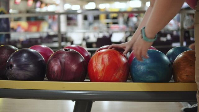 Person spins single ball on rack among many balls in bowling alley. Unidentified woman inserts fingers into the holes of orange ball and picks it up.