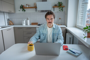 Focused serious man remote working with laptop at kitchen table holding cup coffee, checking plan for day. Morning productivity, balancing work and home comfort, self-discipline, daily work routine.