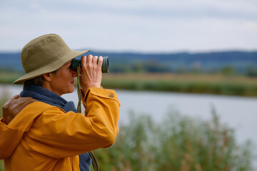 A woman in a yellow raincoat uses binoculars to observe nature by a lake, showcasing a passion for birdwatching and the beauty of the outdoors in a serene setting.