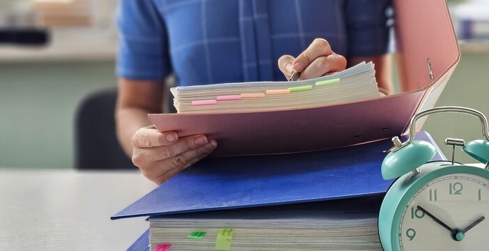 Woman in office with stack of files and folders holds alarm clock