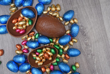 Pile of colourful foil wrapped chocolate easter eggs in pink, blue, gold and green. Large halves of a brown milk chocolate egg have mini eggs inside, on a grey wooden background.