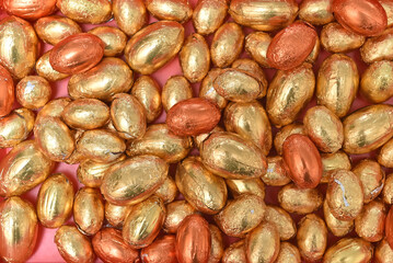Large and small gold, orange and bronze foil wrapped chocolate easter eggs, against a peach orange background.