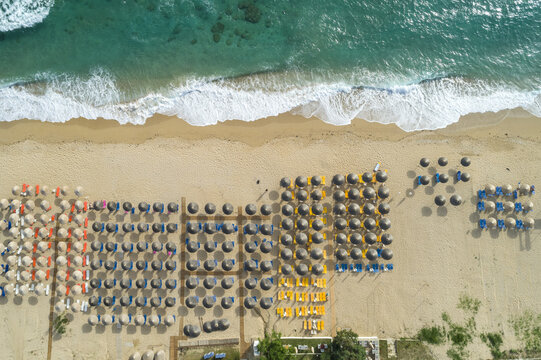 Aerial view of foamy waves caressing the sandy beach with rows of colorful umbrellas and sunbeds creating a vibrant tapestry, Vrachos, Preveza, Greece.