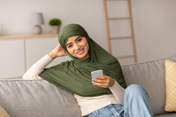 A young Arab woman in a hijab sits comfortably on a sofa at home. She smiles as she uses her smartphone to surf the web and chat online, enjoying her leisure time with family and friends.