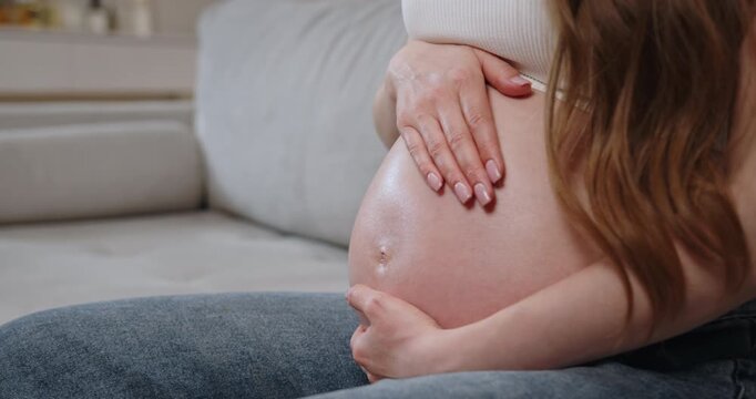Woman's hands gently cradling her pregnant belly while sitting on a couch, showing anticipation and maternal love. Pregnant Belly Close Up