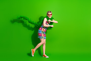 Glamorous young woman in a colorful floral dress runs with a feather boa against a bright green background smiling and showing style and fun