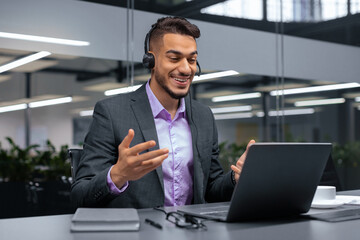 A man in a suit sits at a desk in an office. He is using a laptop and wearing a headset while engaging in a virtual meeting. The setting has modern decor and plants in the background.