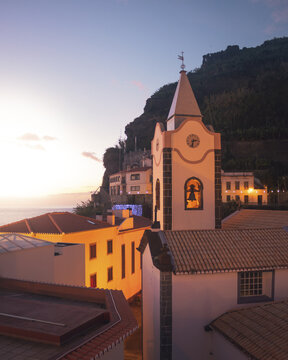 View of warm light bathing the rooftops and church steeple against the dramatic cliffs as dusk settles, Ponta do Sol, Madeira, Portugal.