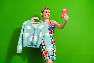 Young glamorous woman poses in colorful floral dress on green screen taking cheerful selfie with a blue daisy cardigan for fashion shopping lifestyle and advertising use