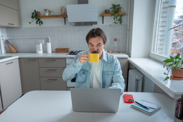 Intently focused man drinking coffee while staring at laptop screen, fully absorbed in work. Concentrated male sipping coffee without looking away from computer. Home office productivity, multitasking