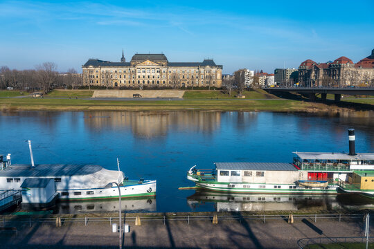 View of the serene Elbe River reflecting the clear sky, with boats docked near the bank and a grand building in the background, Dresden, Saxony, Germany.