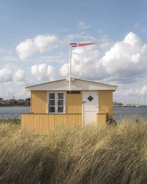 View of a vibrant yellow beach house with a Danish flag fluttering above, nestled in golden grass against a backdrop of a blue sky, Badehuse ved Vestre Strandvej, Ero Municipality, Denmark.
