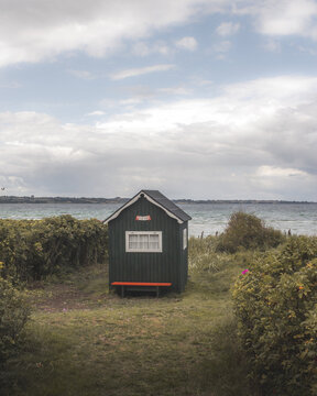 View of a quaint, dark green beach hut with white trim and a vibrant orange bench sits nestled amidst lush greenery near the rippling sea, Badehuse ved Vestre Strandvej, Ero Municipality, Denmark.