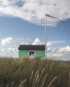 View of a vibrant green beach hut stands proudly amidst golden grasses under a vast, blue sky dotted with fluffy white clouds, Badehuse ved Vestre Strandvej, Ero Municipality, Denmark.