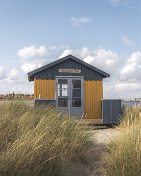 View of a vibrant yellow and blue beach hut stands nestled amongst tall, sun-kissed grasses under a bright sky with scattered clouds, Badehuse ved Vestre Strandvej, Ero Municipality, Denmark.