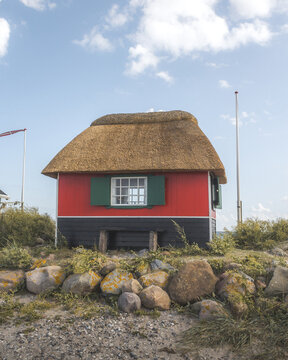 View of a vibrant red and black beach hut with a thatched roof sits atop a rocky outcrop under a serene blue sky, Badehuse ved Vestre Strandvej, Ero Municipality, Denmark.