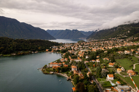 Aerial view of the tranquil lake waters reflecting the overcast sky, contrasting with the vibrant red-roofed houses nestled amongst lush greenery, Lenno, Lombardy, Italy.