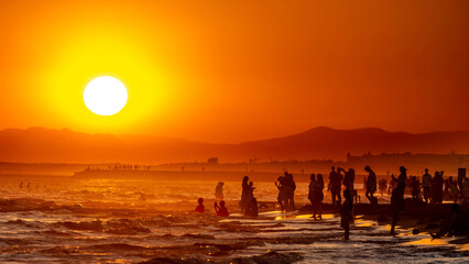 Countless silhouettes of people on a beach against a glowing orange sunset over the shimmering sea. Warm summer vacation atmosphere in backlight. Evrenseki, Turkey, Mediterranean.

