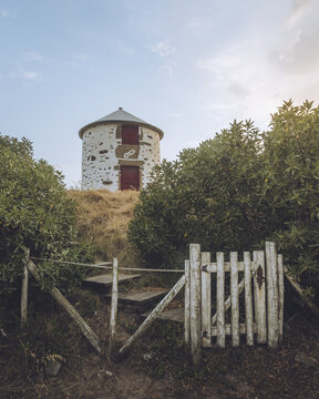 View of a quaint, stone-clad tower with red doors atop a grassy knoll, framed by lush greenery and a weathered wooden gate, Apulia, Braga, Portugal.