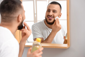 Handsome young happy man cleansing his face with micellar water and cotton pad near mirror in...