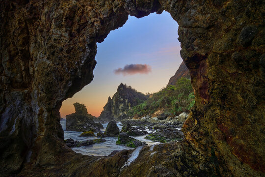 View of the rugged coast through a natural rock window, where the golden sunset kisses the horizon in Watu Lumbung Beach, Yogyakarta, Special Region of Yogyakarta, Indonesia.