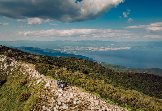 Aerial view of a hiker on a rocky path amidst lush green mountains overlooking the shimmering sea and distant city, Mala Ucka, Primorje-Gorski Kotar County, Croatia.