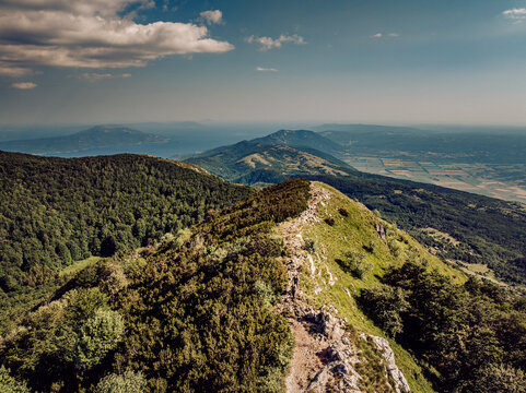 Aerial view of a rugged mountain ridge under a vast sky, where emerald forests meet sun-kissed meadows, Mala Ucka, Primorje-Gorski Kotar County, Croatia.