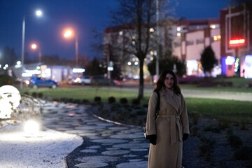 Obraz premium Young woman with long hair wearing a stylish beige coat stands confidently on a city street at night, illuminated by vibrant neon lights, showcasing urban nightlife