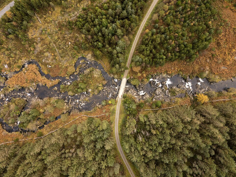 Aerial view of a bridge spanning a dark, rocky river, embraced by autumnal forests in vibrant hues of green, gold, and brown, Newton Stewart, Scotland, United Kingdom.