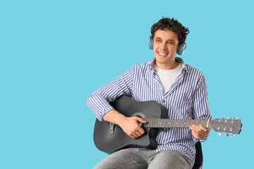 Obraz premium Young happy man in headphones sitting on chair and playing new black guitar against blue background