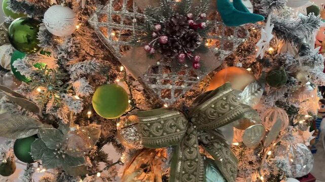 Close up of a Christmas tree with a patterned ribbon bow, star ornaments, pinecones, and glass baubles. Warm lights highlight gold and green holiday decorations in a home interior.